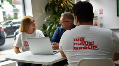 A man in a Big Issue t-shirt talks to a man and a woman at a laptop