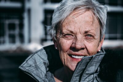 An older woman with short grey hair, wearing a grey coat, smiles at the camera. By Ravi Patel on Unsplash