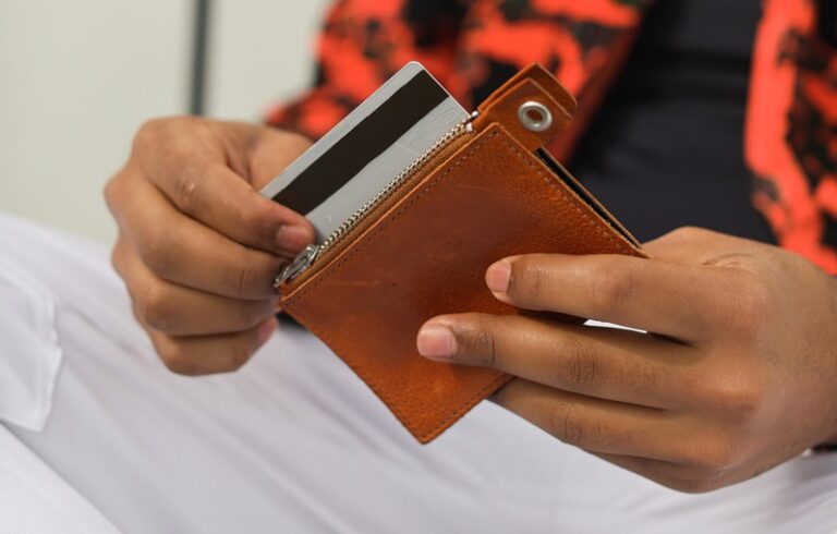 A man's hands take a credit card out of a brown wallet. He is wearing white trousers and a red and black top.