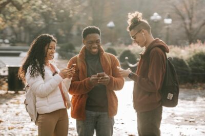 A multiracial group of three young people smile as they look at their phones in a park