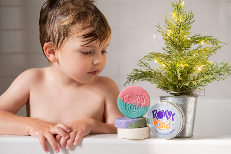 A child leans on the edge of a bath looking at some Rowdy Kind soaps, and next to a small Christmas tree with gold lights