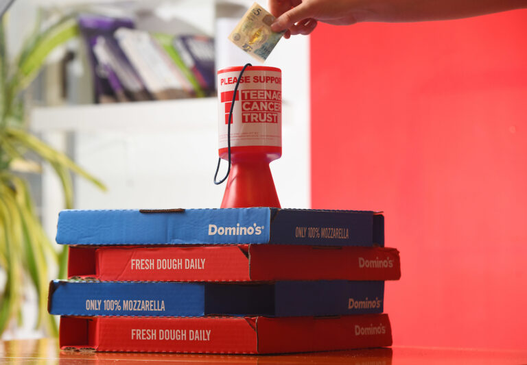 A hand puts note into a Teenage Cancer Trust donation box sitting on a pile of Domino's pizza boxes