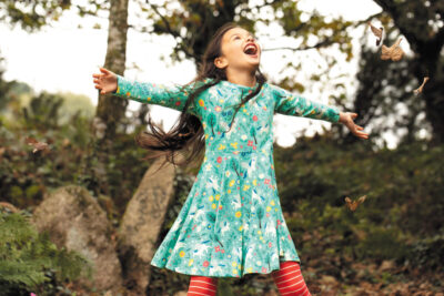 A young girl with long hair in a green patterned Frugi dress holds her arms outstretched and looks up happily at the sky