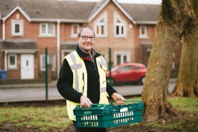 A man in glasses and wearing a hi-vis vest pauses to look at the camera as he carries a green tray of food on a residential street.