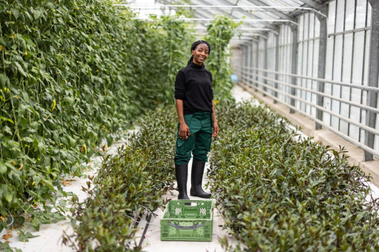 A woman of colour stands facing the camera in a greenhouse. She has green trousers, a black top and wellies on.