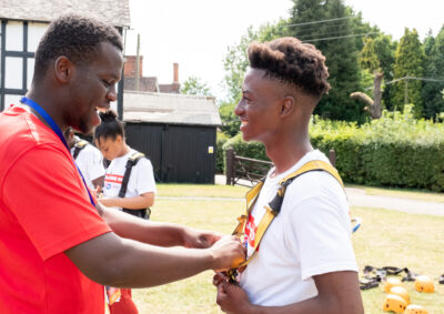 A man helps a smiling teen with a harness for climbing at a StreetGames summer camp