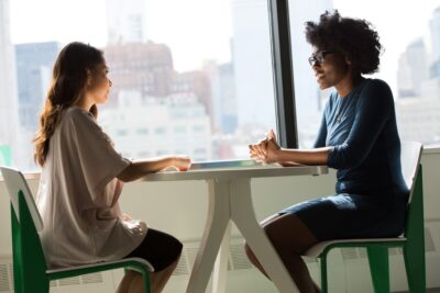 Two women of colour talk across a table in front of a window. By Christina @ wocintechchat.com