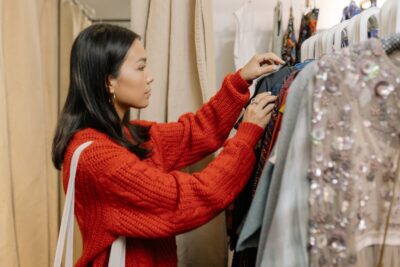 A woman with long dark hair in a reddy-orange jumper looks through clothes on a rack. By Cottonbro on pexels
