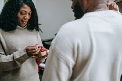 A man looks on as a woman opens a small christmas gift. By Any Lane on Pexels