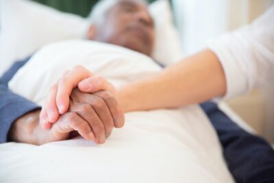 An elderly man lies in a bed, with someone beside him, touching his hand. By kampus production on Pexels