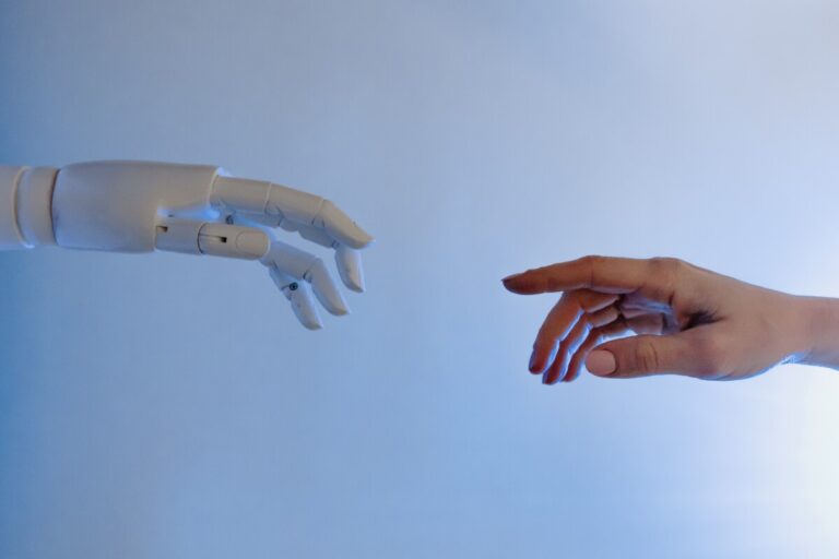 A robot hand and a human hand reach out towards each other against a pale blue background. Photo by Tara Winstead on Pexels