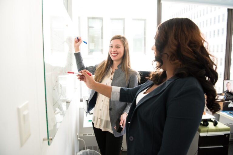 Two women stand at a whiteboard. One is writing and smiling, while the other is facing her and looking away from the camera. By Christina Morillo on Pexels
