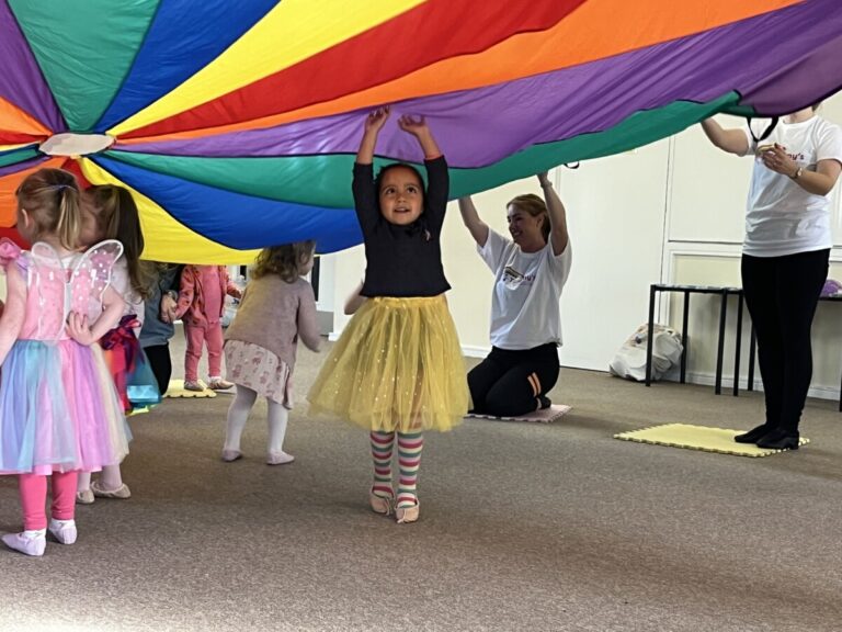 Toddlers dance under a rainbow parachute at a babyballet danceathon