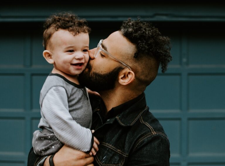 A man kisses his baby son, who is smiling. By Kelly Sikkema on Unsplash