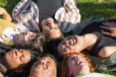 A group of friends lie on the grass with their heads together, laughing. By Kampus Productions on Pexels