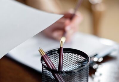 A close up of pens in a pot, with someone signing some papers in the background. By Mikhail Nilov on pexels
