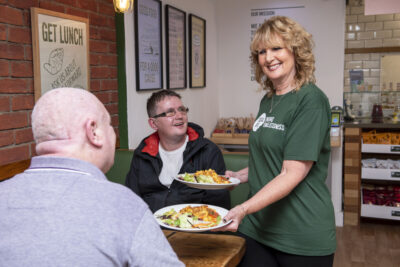 A woman in a dark green charity top smiles as she brings two plates of food to a table where two people are sitting.