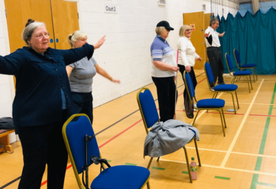People in a hall stand by chairs and do some exercises