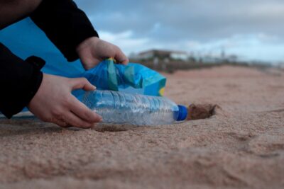 Hands pick up a plastic bottle from a sandy beach and put it in a blue plastic bag. By Marta Ortigosa on Pexels