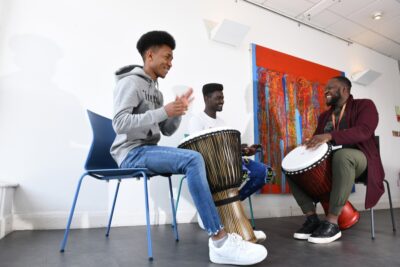 three young Black men laugh and talk as they play drums at the African Community Centre in Wales