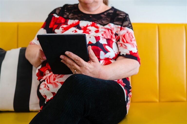 An older woman sits on a yellow sofa, using a tablet. From Good Things Foundation