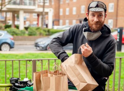 A man puts food into brown paper bags, resting on his bike, outside some blocks of flats