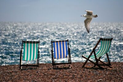 Three deckchairs on a beach with the sea glistening in the background. A gull flies past from the right. Photo: Pexels.com