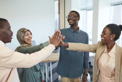 A group of four people of colour high five in a room with tables and a big window. By Edmond Dantes on Pexels