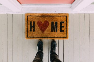 Looking down at a welcome mat that says Home with a red heart replacing the O. By Kelly on Pexels