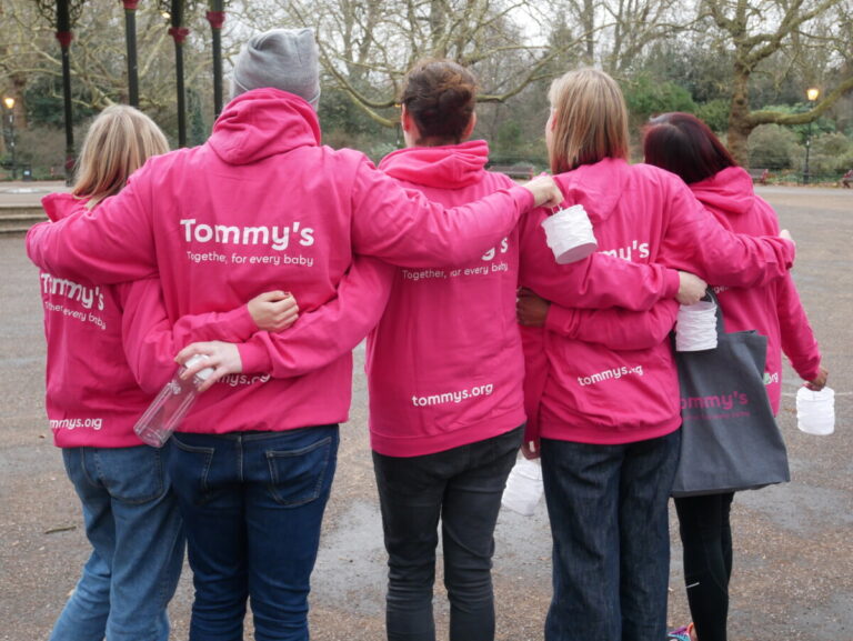 5 people link arms with their backs to the camera, dressed in pink Tommy's Walk for Hope hoodies