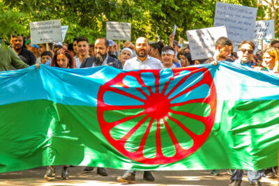A group of Roma people protesting, holding up the flag of the Roma with is blue and green with a red dharmachakra, or cartwheel in the centre