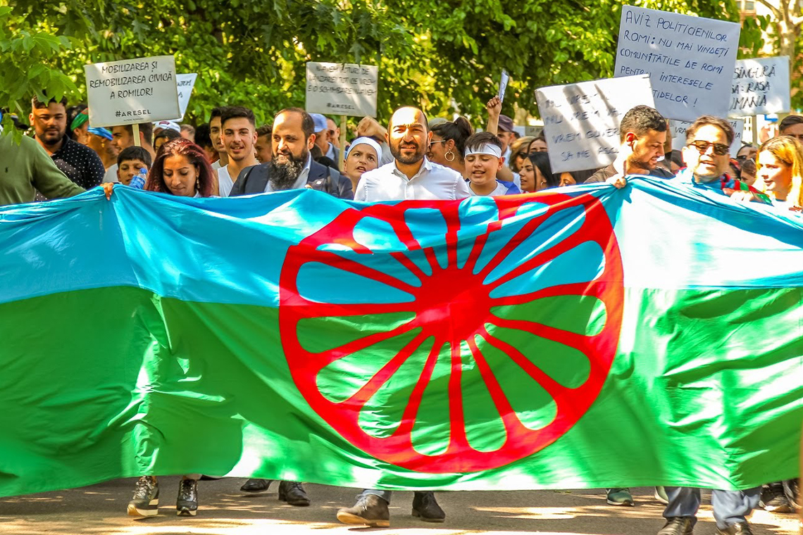 A group of Roma people protesting, holding up the flag of the Roma with is blue and green with a red dharmachakra, or cartwheel in the centre