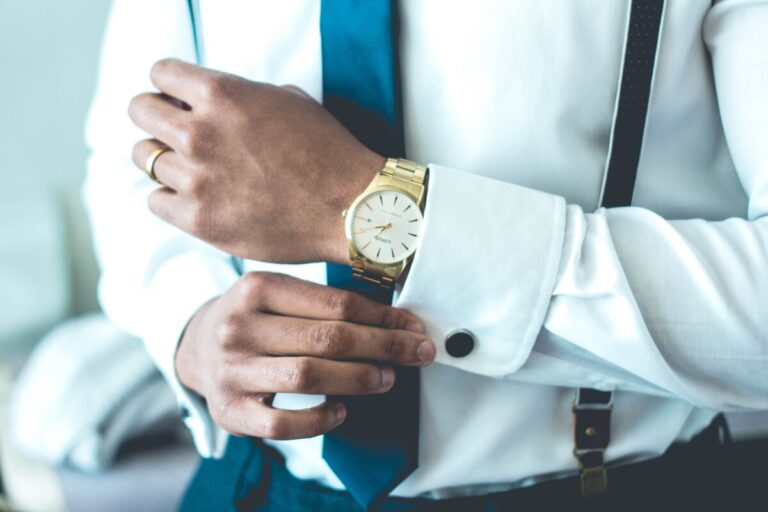 A man's hand adjusting his cufflink on a white shirt, with a blue tie, and a gold watch. By Jonathan Francisca on Unsplash.