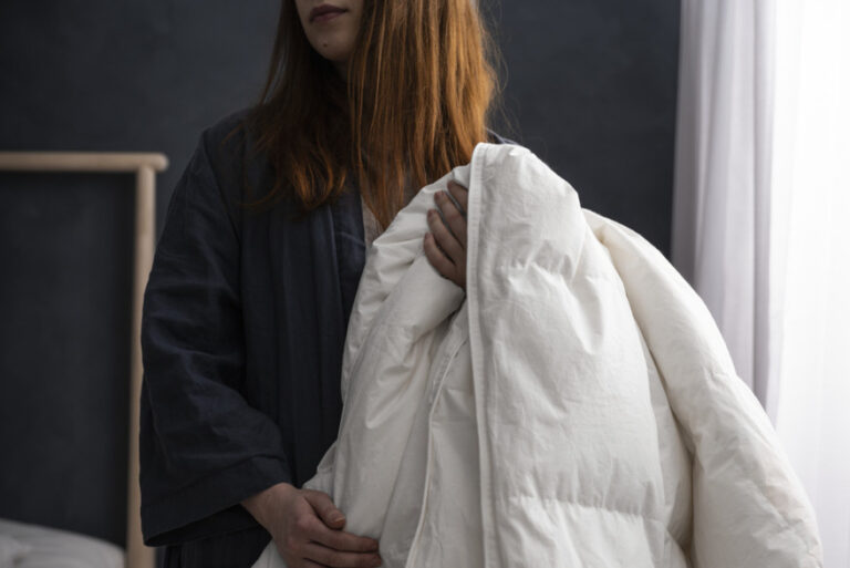 A young woman holds a duvet by the side of a bed