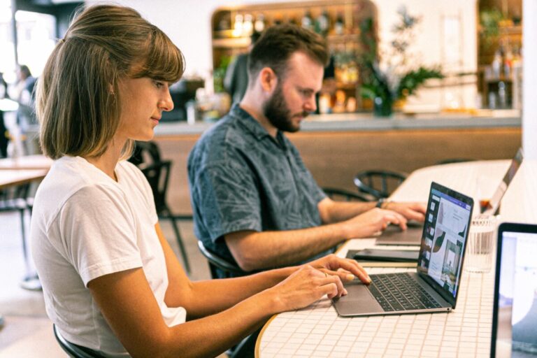 two people, a man and a woman sit side by side at a table, working on their laptops. By Canva Studio on Pexels