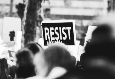 A black and white photo of a sign saying resist, held up at a protest. By Sides Imagery on Pexels