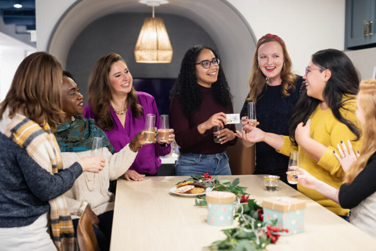 Women celebrating Christmas with drinks and mince pies stand around a table, looking happy. By Lisa Damico Portraits