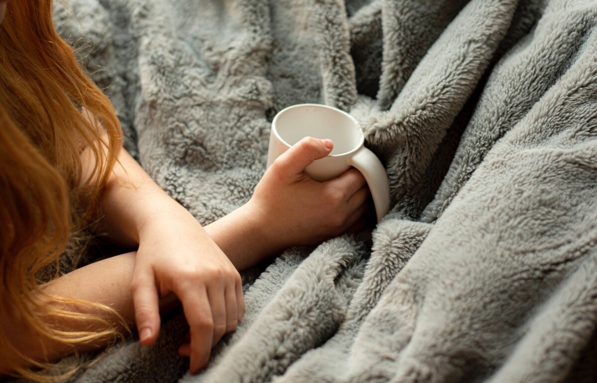 A woman's arms on top of a grey blanket, in one hand she holds a white mug. By Amin Hasani on Unsplash