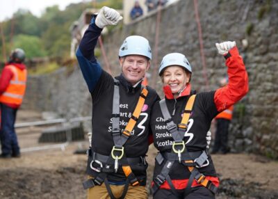 A man and a woman i climbing gear raise their fists triumphantly after completing the Forth Bridge Abseil