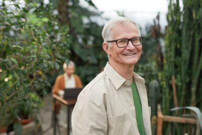An older man with short white hair and black framed glasses smiles at the camera from a garden. There is a woman in the background. By Marcus Aurelius on Pexels
