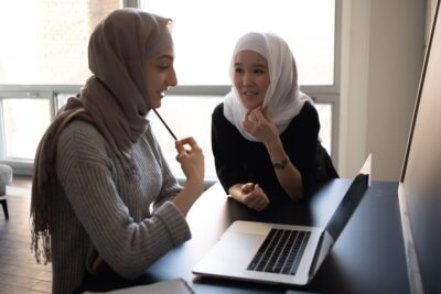 Two young Muslim women wearing hijabs talk over a laptop. By Monstera Production on pexels