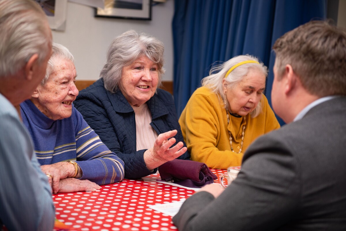 A group of older people sit chatting around a table with red checked tablecloth. From Age Sdotland