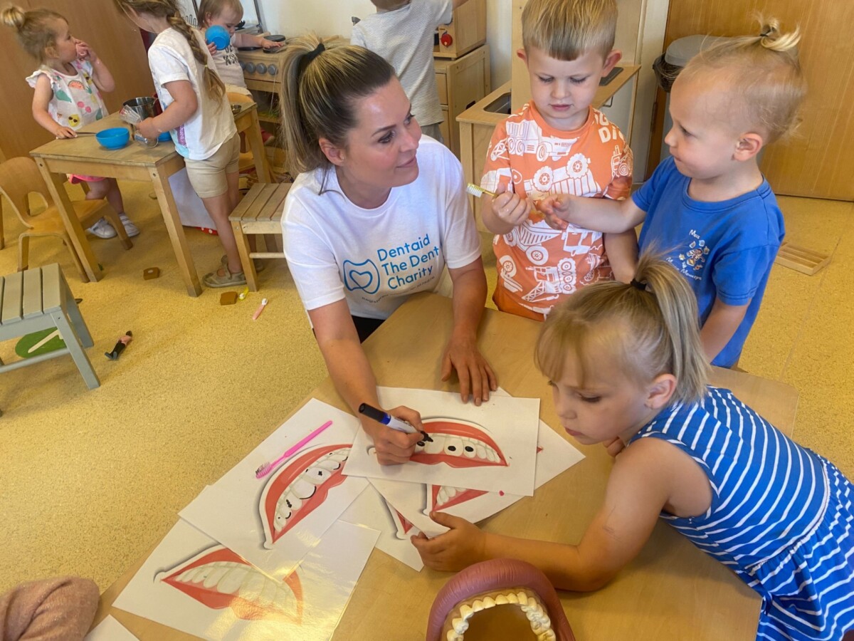A Dentaid staff-member joins three young children at a nursery where they are colouring in pictures of mouths and teeth.