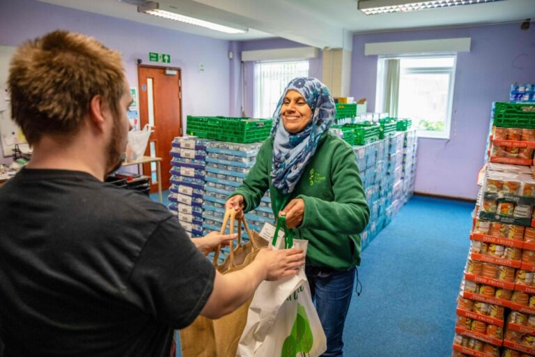 A smiling Trussell Trust volunteer hands over a bag of food items to a young man.