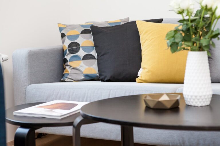 A photo of yellow and grey cushions on a light grey sofa. behind a coffee table with a vase. By Terje Sollie on Pexels
