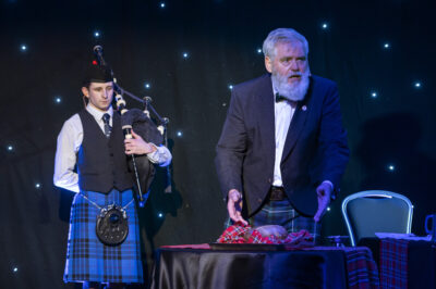 A bagpiper looks on as a bearded man addresses a haggis for Burns Night