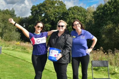 Three women on a sunny day pose with a collecting bucket for Cancer Research UK