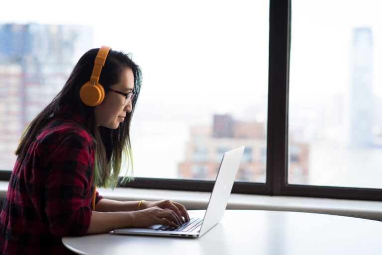 A woman with long dark hair, glasses and orange headphones types on her laptop. By Christina Morillo on pexels