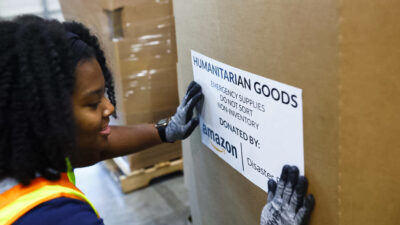 A young woman puts an Amazon label on a cardboard box saying 'humanitarian goods'.
