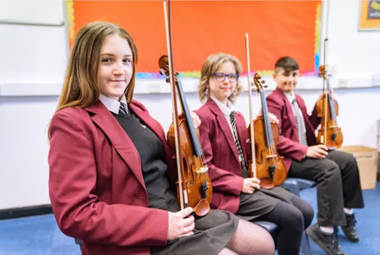 Three teenagers in school uniform hold their violins and look at the camera.